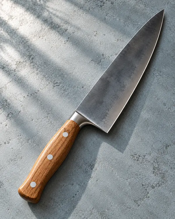 Long carbon-steel chef's knife with an oak handle on a muted blue-grey surface